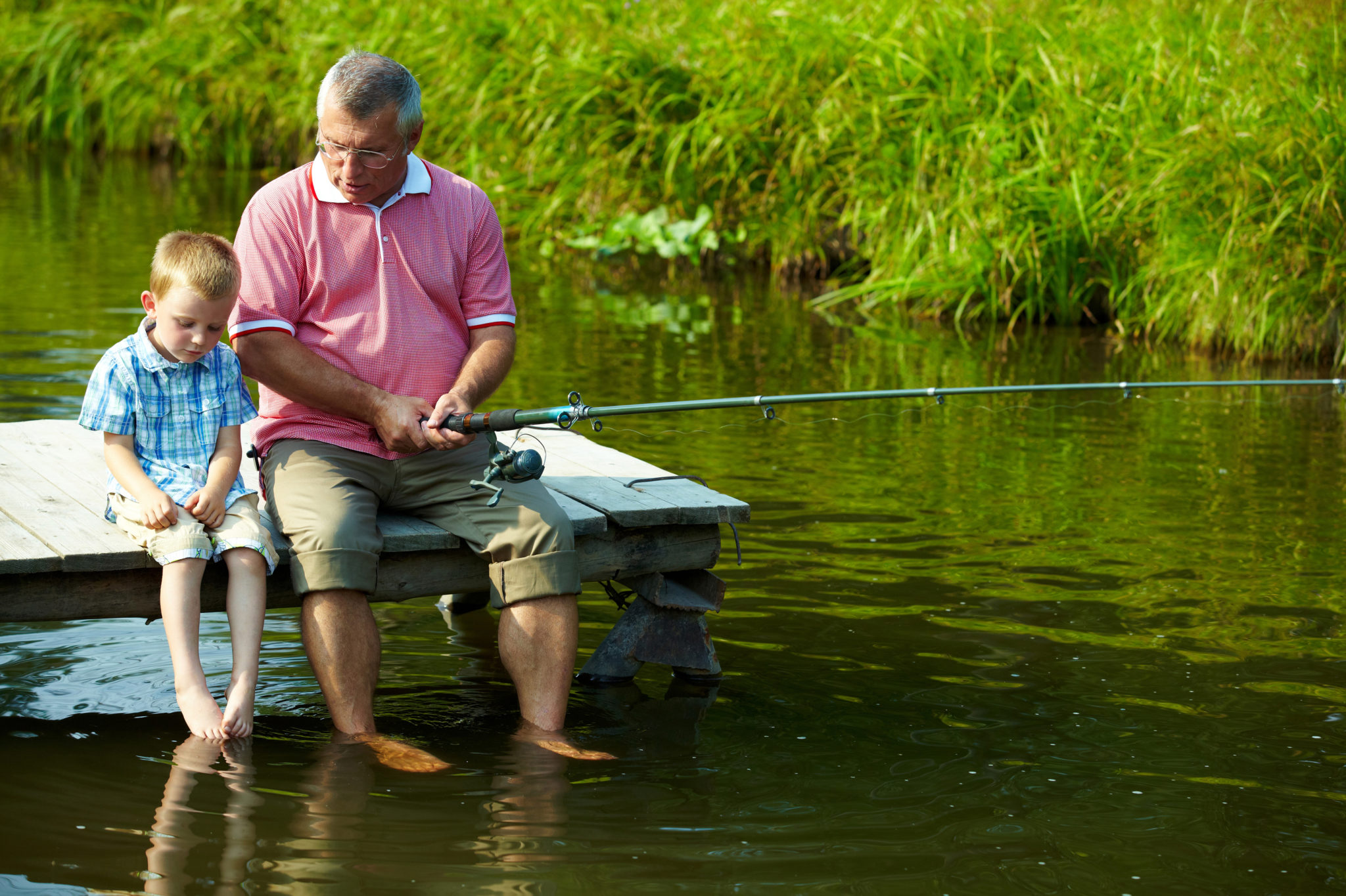 Photo of grandfather and grandson sitting on pontoon with their feet in water and fishing on weekend